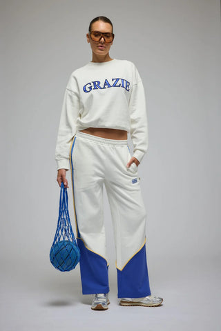 Person wearing a white 'Grazie' sweatshirt and white and blue pants, holding a blue bag on a gray background.